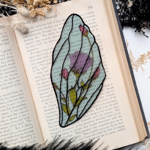 May include: A stained glass butterfly wing bookmark with pink and green floral details, resting on an open book. The book's pages display text from "Pit and the Pendulum" by Edgar Allan Poe.