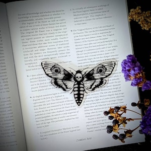May include: A black and white moth sticker with a skull design, placed on an open book. The book's pages display text, and dried flowers and berries are arranged nearby, creating a gothic aesthetic.