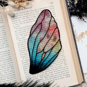 May include: A colorful stained glass butterfly bookmark with a black outline. The butterfly has a rainbow gradient of colors, including pink, orange, yellow, green, blue, and purple. The bookmark is placed on a page of a book with text about "Pit and the Pendulum".