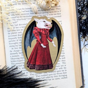 May include: A sticker featuring a wolf-like creature in a red dress with black bat wings. The illustration has a vintage aesthetic, with the creature's face showing sharp teeth. The sticker is set against a gold background.
