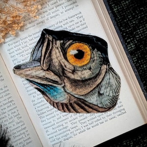 May include: A die-cut owl eye sticker with a yellow iris and black and white details, placed on an open book. The sticker is a unique decorative item, perfect for book lovers and those who appreciate whimsical art.