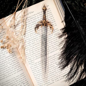 May include: A decorative bookmark featuring a small, ornate sword design. The bookmark is placed on an open book with dried flowers and a black feather, creating a gothic aesthetic.