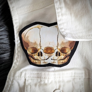 May include: A black and white patch with an image of two conjoined skulls. The skulls are detailed and have a realistic appearance.