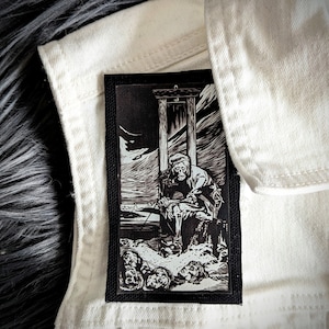 May include: Black and white embroidered patch featuring a skeletal figure sitting on a platform with a guillotine above. The figure is wearing a hat and holding a staff.