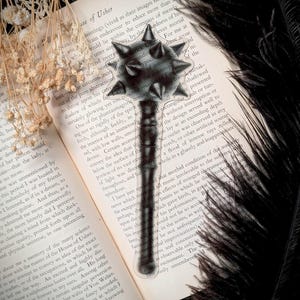 May include: A clear, transparent bookmark shaped like a medieval mace, with a spiked head and a long handle. The bookmark is placed on an open book with text, surrounded by black feathers and dried flowers.