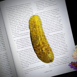 May include: A yellow pickle bookmark with a textured surface. The bookmark is partially covering a page of a book with text about the legend of Dr. Faust.