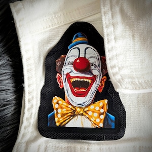 May include: A black and white patch with a colorful image of a clown with a red nose and a yellow bow tie. The clown is smiling and has a large, open mouth.