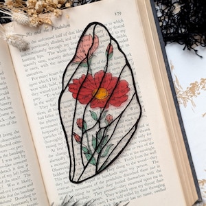 May include: A floral bookmark featuring a red flower with a yellow center and green leaves, outlined in black. The bookmark is placed on an open book with text visible. Decorative elements include dried flowers and black feathers.