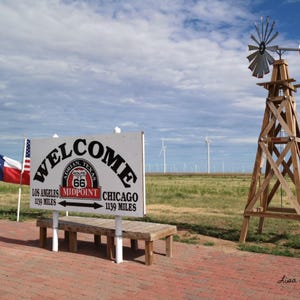 Op de afbeelding: Een wit bord met de tekst "WELCOME" in een rode boog, met de woorden "ADRIAN TEXAS" en "ROUTE 66" in kleinere tekst eronder. Het bord zegt ook "LOS ANGELES 1139 MILES" en "CHICAGO 1139 MILES" met pijlen die naar links en rechts wijzen. Het bord staat voor een houten bank en een houten windmolen met een Texasvlag.