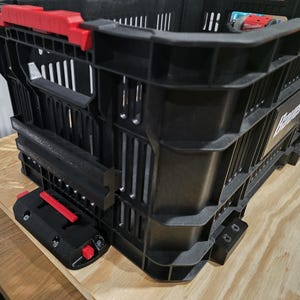 May include: Black plastic storage crate with red accents on the handles and a side-mounted accessory. The crate is sitting on a wooden surface. The accessory has a red lever.