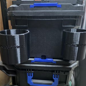 May include: Black 3D-printed cup holders with the text "DKolo3DPrintz" are displayed in front of a black toolbox. The toolbox has blue handles and latches. The cup holders are cylindrical and appear to be designed to attach to the toolbox.