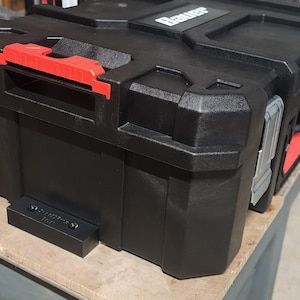 May include: Black plastic tool box with a red handle and silver latches. The box has a textured surface and the brand name "Bauer" is visible on the top. The box is sitting on a wooden surface.