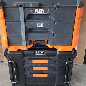 May include: Two stacked toolboxes, one black and orange with the Klein Tools logo, and the other black with the Ridgid logo. The top toolbox has three drawers and a gray strap. The bottom toolbox has a hinged lid.