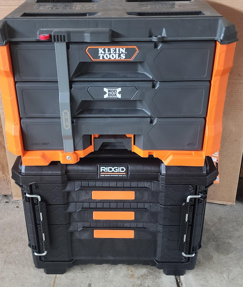 May include: Two stacked toolboxes, one black and orange with the Klein Tools logo, and the other black with the Ridgid logo. The top toolbox has three drawers and a gray strap. The bottom toolbox has a hinged lid.