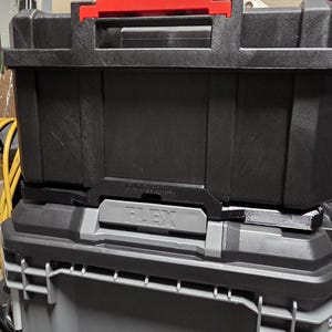 May include: Black and gray toolboxes stacked on top of each other. The top toolbox has a red handle and the word "FLEX" is visible on the front of the bottom toolbox. The toolboxes are made of a durable plastic material.