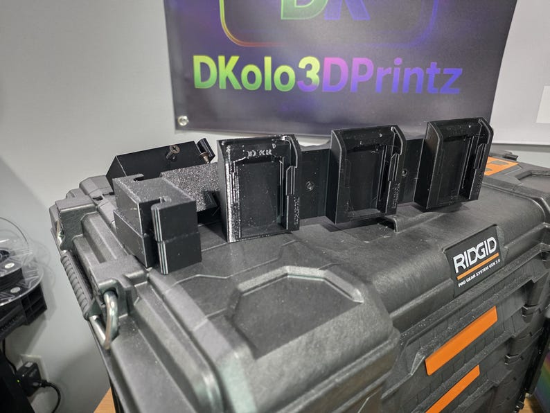 以下が含まれることがあります： 黒色の3Dプリントされたツールホルダーが、黒色のRidgid Pro Gear System 2.0ツールボックスに取り付けられています。 ホルダーはツールを安全に保持するように設計されています。 背景のバナーには「DKolo3DPrintz」と書かれています。