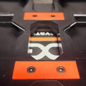 May include: Close-up of a black plastic object with orange accents. Two screws are visible on an orange rectangular piece. The object has a recessed area with the letters "DX" and the word "VST" visible.