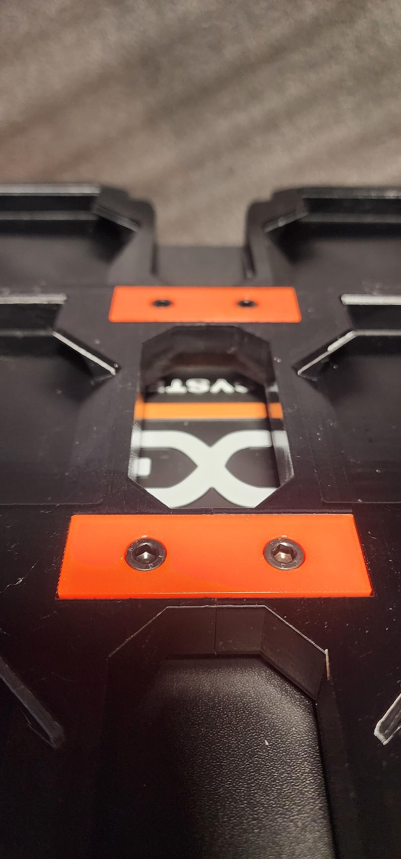 May include: Close-up of a black plastic object with orange accents. Two screws are visible on an orange rectangular piece. The object has a recessed area with the letters "DX" and the word "VST" visible.