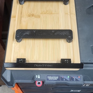 May include: A bamboo cutting board with black 3D-printed brackets attached. The brackets have a textured carbon fiber appearance. The text "DKolo3DPrintz" is visible on one of the brackets. The board is placed on a gray and black tool box.