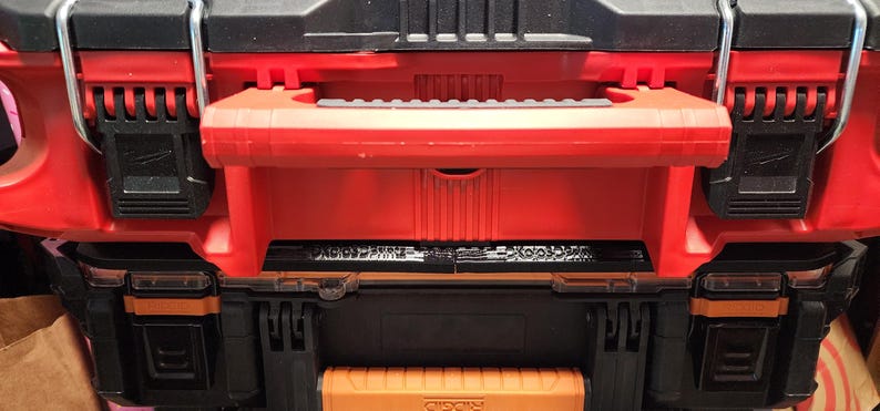 May include: A red and black tool box with a handle and multiple latches. The tool box has a black top and bottom section with a red center section. The latches are black and the handle is red.
