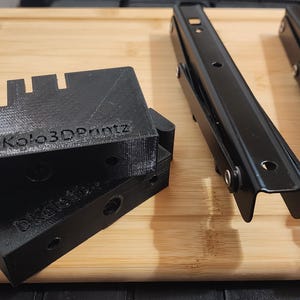 May include: Black metal and 3D printed components, including brackets and hinges, are arranged on a wooden surface. The 3D printed parts have the text "DKolo3DPrintz" on them. The components appear to be for a mechanical or structural application.