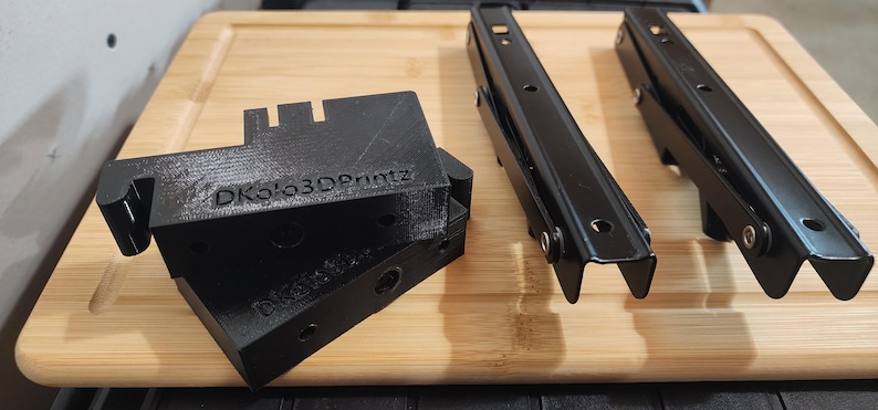 May include: Black metal and 3D printed components, including brackets and hinges, are arranged on a wooden surface. The 3D printed parts have the text "DKolo3DPrintz" on them. The components appear to be for a mechanical or structural application.