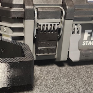 May include: Close-up of a black and gray FLEX Stack Pack tool case. The case features a black latch and a black and gray bracket. The FLEX logo is visible on the side of the case. The case is sitting on a gray surface.