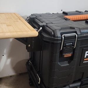 May include: A black RIDGID Pro Gear System 2.0 tool box with an attached light-colored wooden cutting board. The cutting board is rectangular and has a recessed edge. The tool box has orange accents and metal latches.