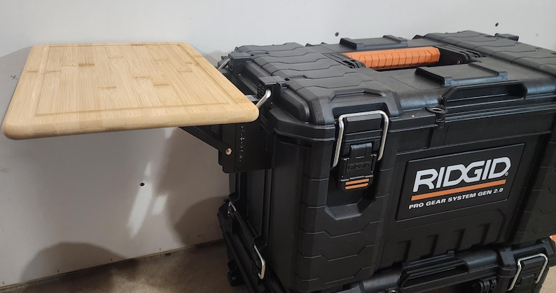 May include: A black RIDGID Pro Gear System 2.0 tool box with an attached light-colored wooden cutting board. The cutting board is rectangular and has a recessed edge. The tool box has orange accents and metal latches.