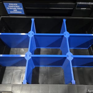 May include: A black tool storage drawer featuring a blue plastic grid divider system that creates six separate compartments. A blue rectangular label above the drawer displays the white text: PADLOCK COMPATIBLE TO SECURE TOOLS AND ACCESSORIES.