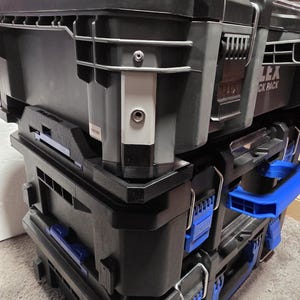 May include: Black modular tool storage system with blue accents. The system is composed of several stacked units, each with secure latches and handles. The word "FLEX" is visible on the top unit. The system appears durable and designed for organization.