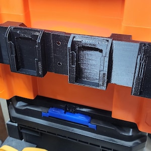 May include: A black 3D-printed plastic battery mount rack featuring four individual slots for power tool batteries. The rectangular mounting bar has visible screw holes and is attached to an orange storage case with a textured surface finish.