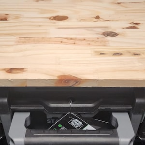 May include: A light-colored wooden top sits atop a black and gray storage container. The wood grain is visible, with knots and variations in color. The container has metal latches and a handle. A tag hangs from the container.