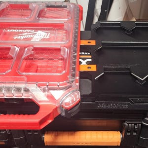 May include: Two interlocking toolboxes. One is red with a clear lid and the word "PACKOUT" and "Milwaukee" visible. The other is black with orange accents and the word "MILWAUKEE" visible. Both have handles and are designed for tool storage.