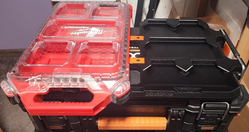 May include: Two interlocking toolboxes. One is red with a clear lid and the word "PACKOUT" and "Milwaukee" visible. The other is black with orange accents and the word "MILWAUKEE" visible. Both have handles and are designed for tool storage.