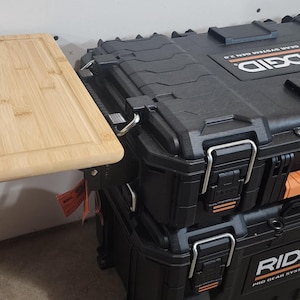 May include: A light-colored bamboo cutting board is attached to a black RIDGID Pro Gear System Gen 2.0 tool storage box. The box has orange accents and metal latches. The cutting board is rectangular.