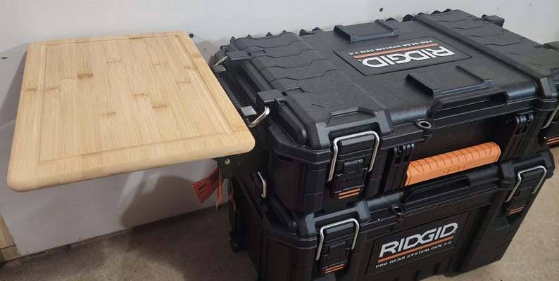 May include: A light-colored bamboo cutting board is attached to a black RIDGID Pro Gear System Gen 2.0 tool storage box. The box has orange accents and metal latches. The cutting board is rectangular.