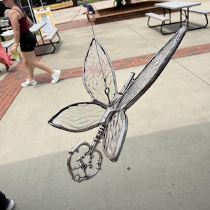 May include: A hanging ornament shaped like a dragonfly with clear, textured glass wings framed in dark metal. The body of the dragonfly is formed by a silver-toned metal skeleton key with an ornate, circular bow and a long, slender shaft.