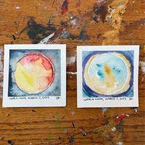 May include: Two watercolor paintings of the Worm Moon, a full moon in March. The first painting is a red and yellow moon on a blue background. The second painting is a blue and yellow moon on a purple background. Both paintings have the text "WORM MOON, MARCH 7, 2023" written below them.