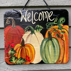 May include: A black slate sign with the word "Welcome" painted in white. The sign is decorated with painted pumpkins in shades of orange, green, white, and red.