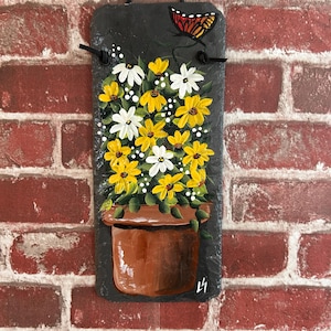 May include: Hand-painted slate wall hanging featuring a brown pot filled with yellow and white flowers. A butterfly is painted in the upper right corner. The slate is hung with a black cord. The background is a red brick wall.
