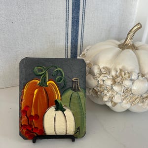 May include: A rectangular slate plaque featuring a hand-painted design of three pumpkins in orange, white, and green, accented with red berries and green vines. The artwork is displayed on a small black stand, showcasing a signature in the bottom corner.