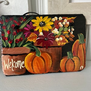 May include: Hand-painted welcome sign on a black slate. The sign features a floral arrangement with sunflowers, burgundy daisies, and berries in brown pots. Orange pumpkins are painted on the right side, and the word "Welcome" is written in white.