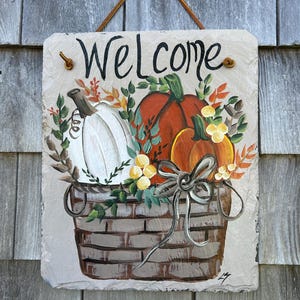 May include: A rectangular slate sign with a hanging leather cord, featuring a hand-painted basket filled with a white pumpkin, an orange pumpkin, and autumn foliage. The word "Welcome" is written in black script at the top of the sign.