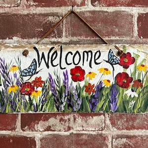 May include: A painted slate welcome sign with a floral design. The sign features a butterfly, red, yellow, and purple flowers, and the word "Welcome" in black lettering.