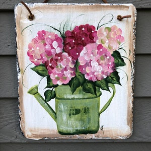 May include: Hand-painted slate sign featuring a green watering can filled with pink and burgundy hydrangeas. The sign has a natural edge and a leather strap for hanging. The background is off-white with a brown border.