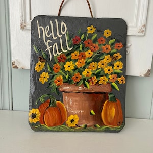 May include: A rectangular slate sign with a painted fall theme. The sign features a brown pot filled with yellow and orange flowers, two orange pumpkins, and the words "hello fall" in white script. A brown leather strap is attached for hanging.