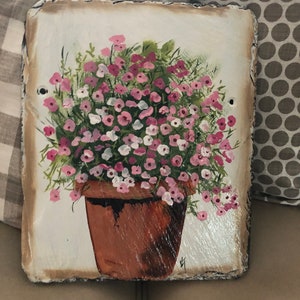 May include: Hand-painted floral art on a square slate tile. The artwork features a brown terracotta pot filled with pink and white flowers and green foliage. The tile has a rough, textured edge and is set against a neutral background.