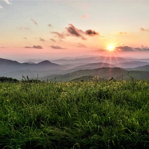 May include: A scenic landscape photograph of a mountain range at sunset. The sky transitions from pink to orange, with the sun's rays bursting through the clouds. Lush green grass and wildflowers fill the foreground.