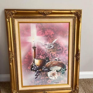 May include: A framed painting depicting a religious scene with a white dove, a chalice, bread, grapes, and a candle in front of a cross. The painting has a pink and purple background with a gold frame.